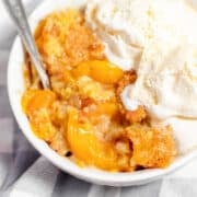 A bowl of peach dessert with baked peaches and crust, reminiscent of a peach dump cake, topped with two scoops of vanilla ice cream and a spoon, on a checkered cloth.