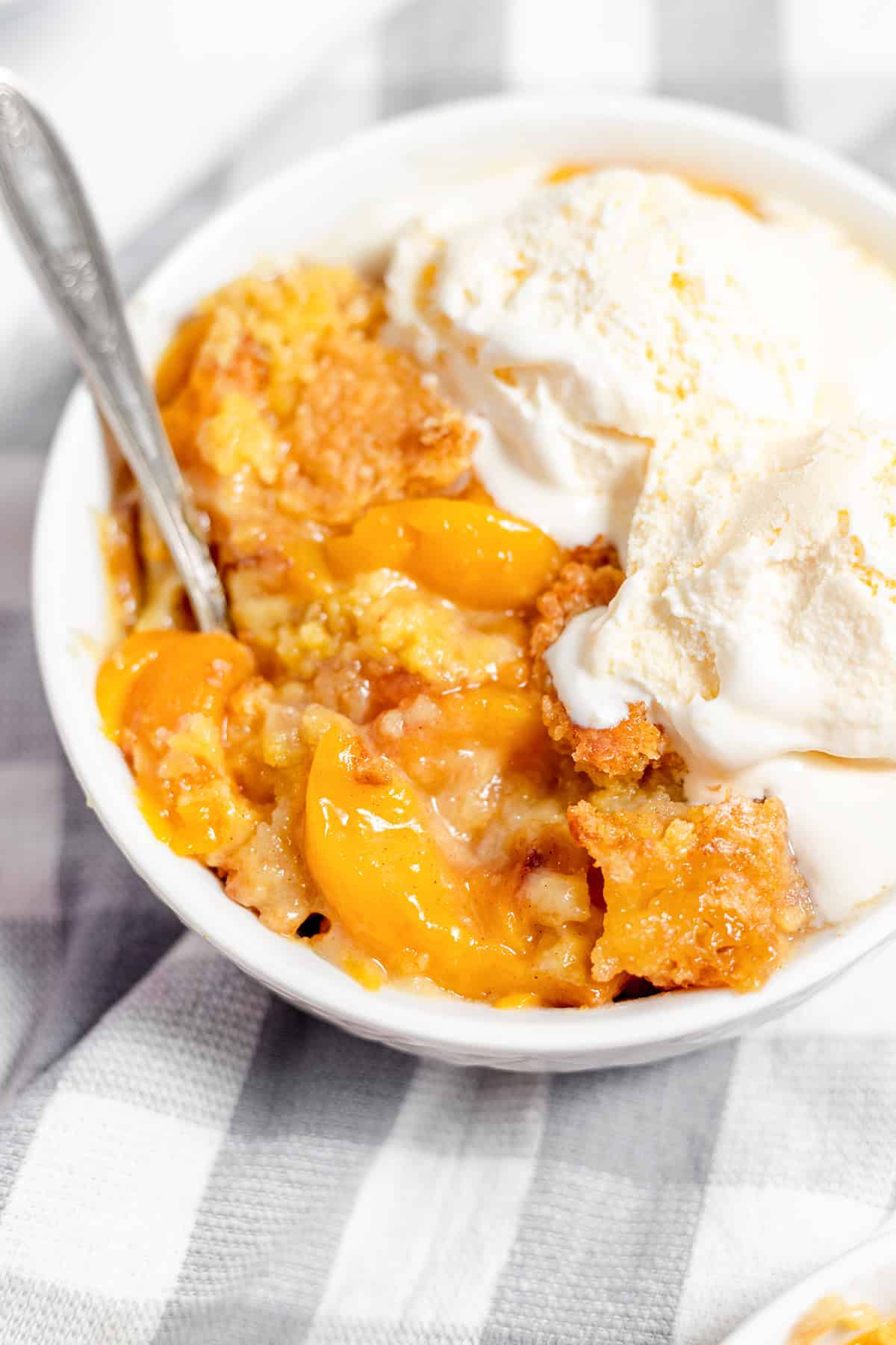 A bowl of peach dessert with baked peaches and crust, reminiscent of a peach dump cake, topped with two scoops of vanilla ice cream and a spoon, on a checkered cloth.