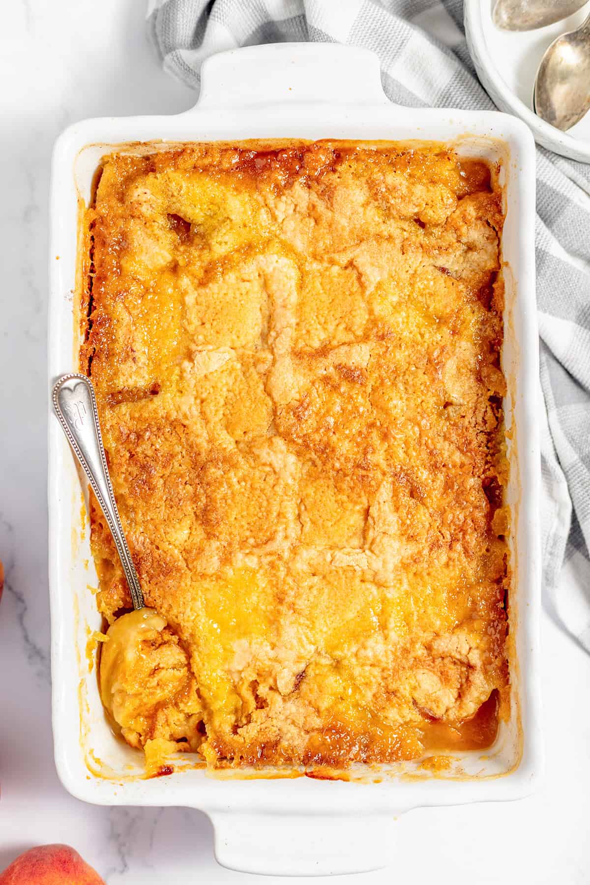 A rectangular white baking dish filled with golden-brown Peach Dump Cake, with a serving spoon scooping out a portion from one corner.