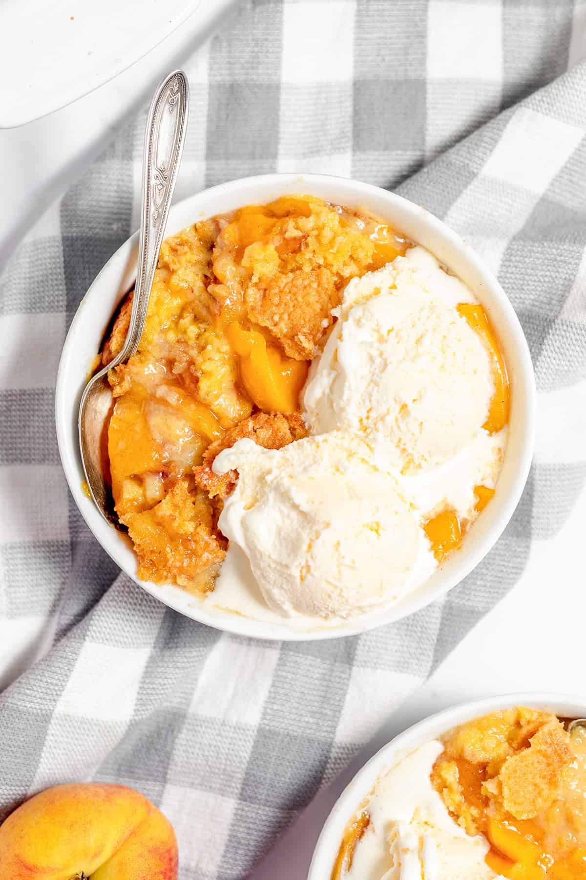 A bowl of warm peach cobbler, a classic peach dessert, topped with two scoops of vanilla ice cream and served with a spoon on a gray and white checkered cloth.