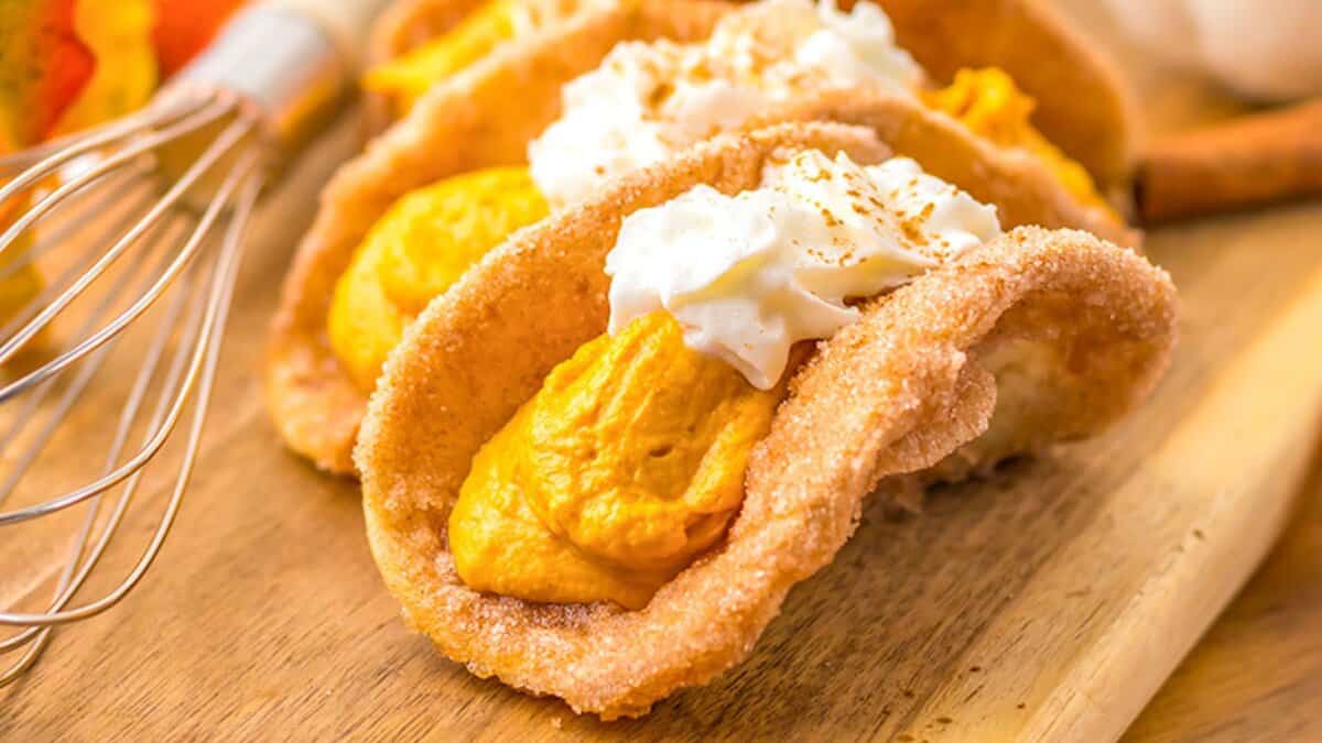 Close-up of dessert tacos with cinnamon-sugar shells, filled with pumpkin mousse and topped with whipped cream—a perfect addition to your collection of Autumn Desserts—displayed on a wooden surface.