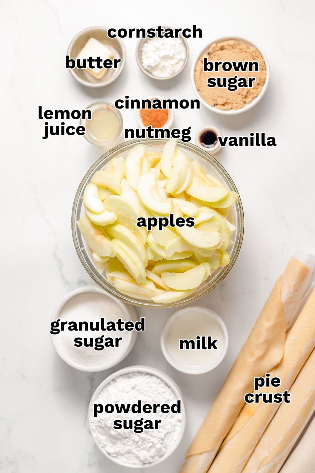 Labeled ingredients for Apple Slab Pie filling and crust are arranged on a white surface, including sliced apples, sugars, spices, corn starch, butter, lemon juice, vanilla, milk, and rolled pie crust.
