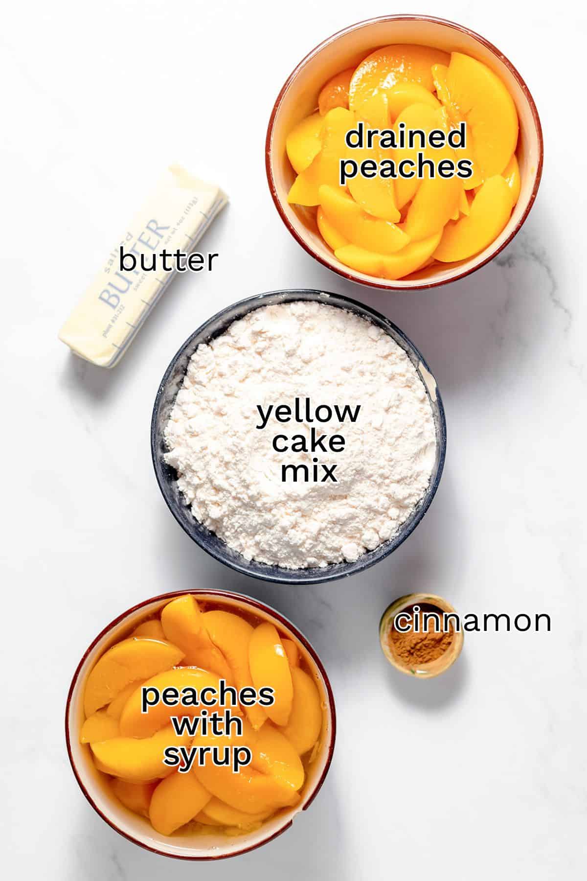 Overhead view of ingredients for Peach Dump Cake: butter, yellow cake mix, cinnamon, a bowl of drained peaches, and a bowl of peaches with syrup arranged on a white surface—perfect for an easy dessert.