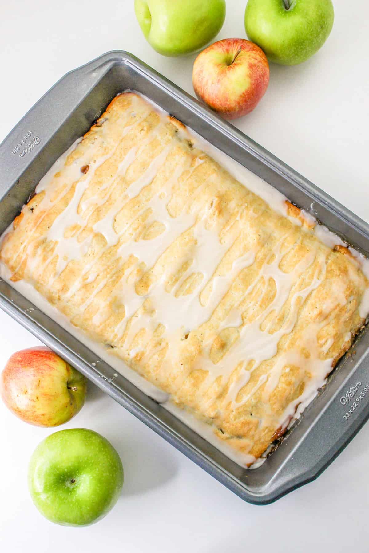 A rectangular baked dessert bar with icing sits in a metal pan, surrounded by green and red apples on a white surface—perfect for fans of Apple Pie Bars.