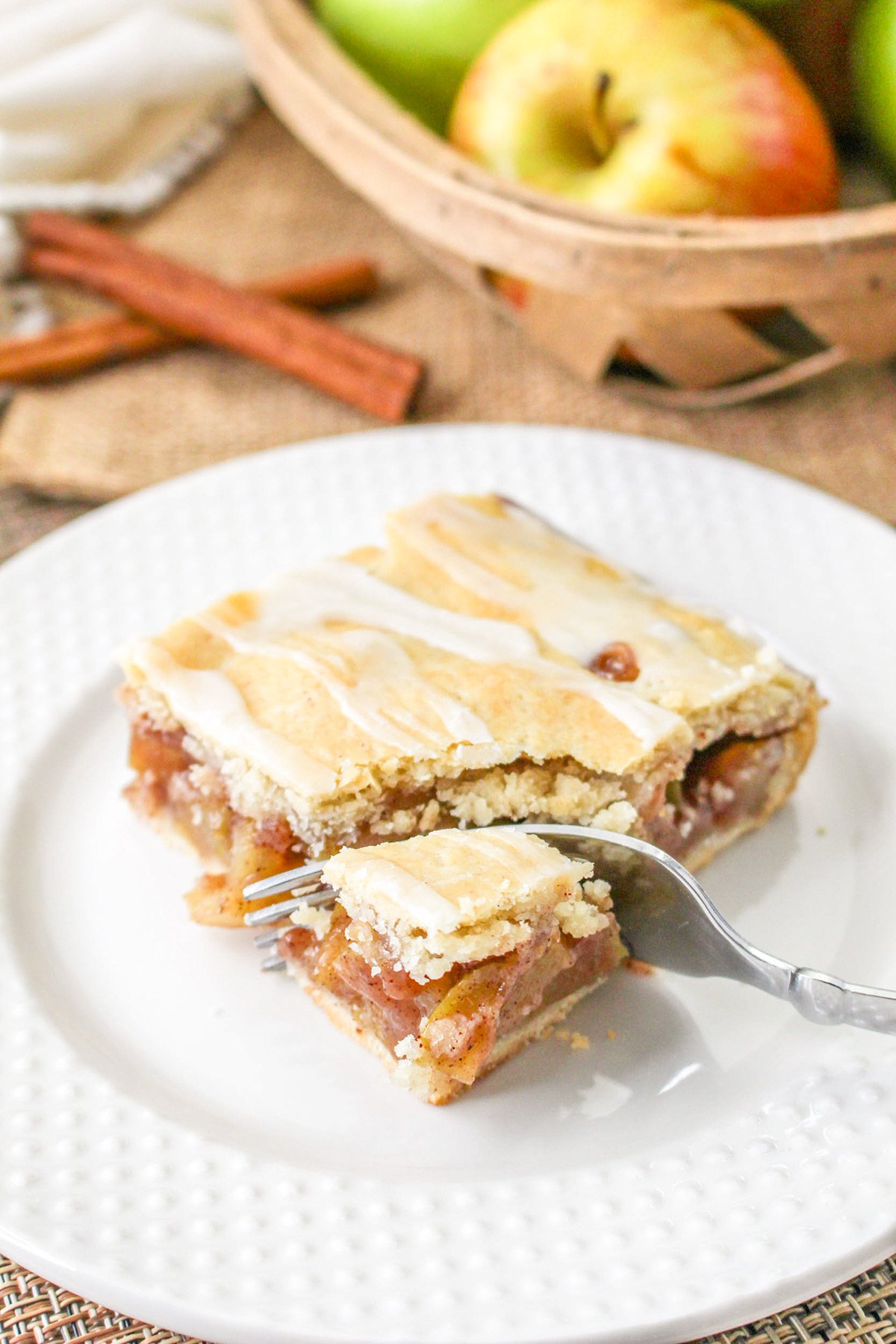 A slice of Apple Pie Bars with a sweet glaze sits on a white plate beside a fork; apples and cinnamon sticks add a touch of warmth in the background.