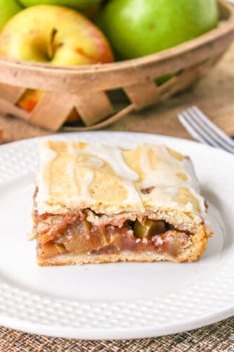 A slice of Apple Pie Bars sits on a white plate, with a basket of apples in the background.