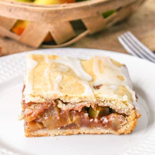 A slice of Apple Pie Bars sits on a white plate, with a basket of apples in the background.