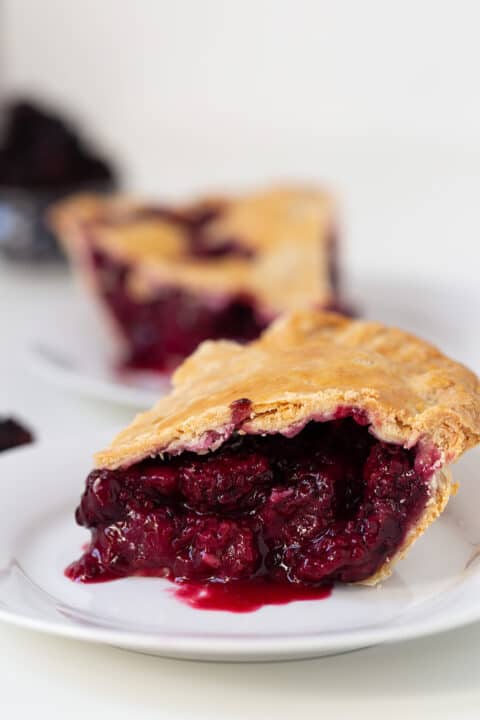 Two slices of blackberry pie with golden crust and rich, dark berry filling are served on white plates.
