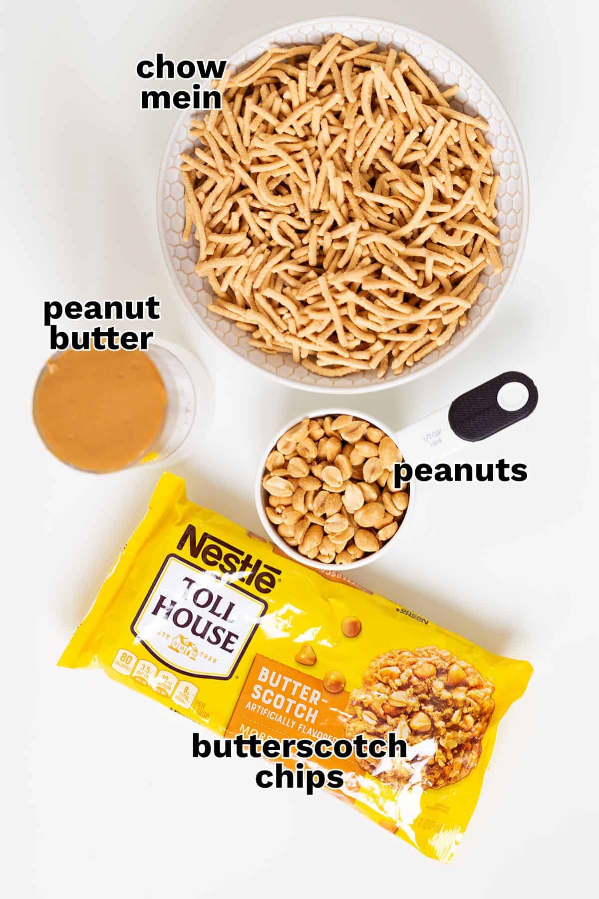 A bowl of chow mein noodles, a glass of peanut butter, a cup of peanuts with a measuring spoon, and a package of Nestlé butterscotch chips set on a white background—classic ingredients for Butterscotch Haystacks, an easy no bake dessert.