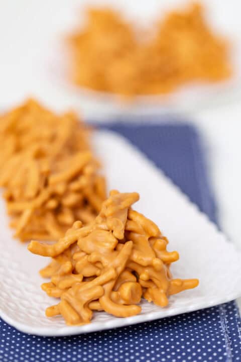 Close-up of No Bake Butterscotch Haystacks cookies arranged on a white plate, with more butterscotch cookies blurred in the background.