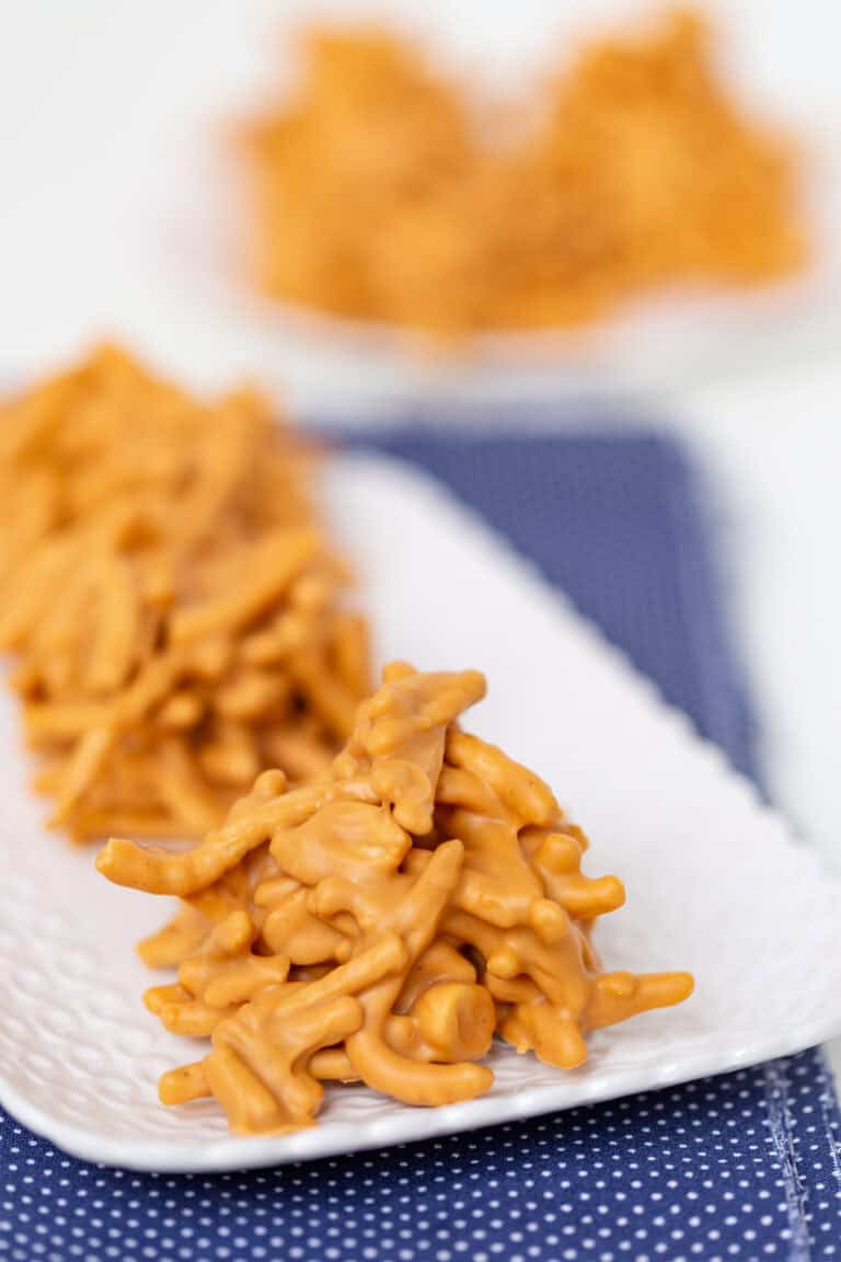 Close-up of No Bake Butterscotch Haystacks cookies arranged on a white plate, with more butterscotch cookies blurred in the background.