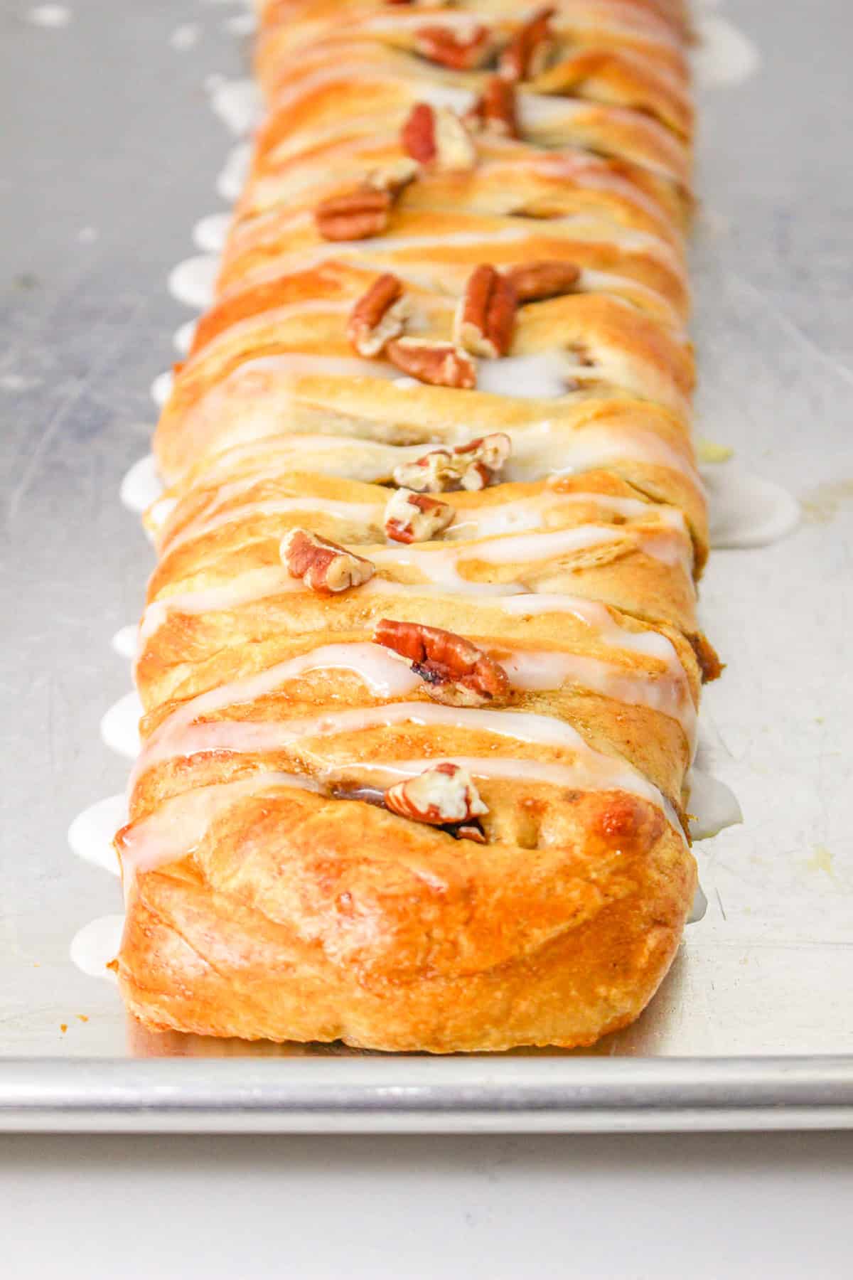 A Crescent Roll Braid filled with pumpkin and topped with icing and pecan halves is displayed on a metal baking sheet.