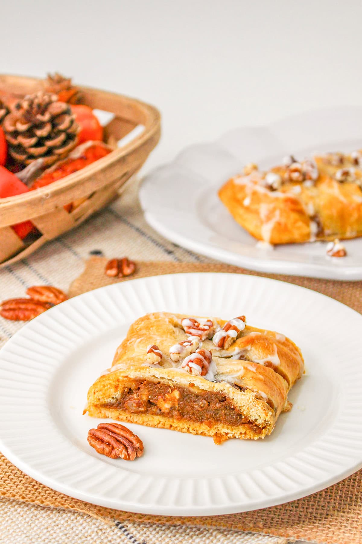 A slice of pecan braid pastry with icing and pecans on a white plate, with a full pastry and a basket of pinecones and fruit in the background.