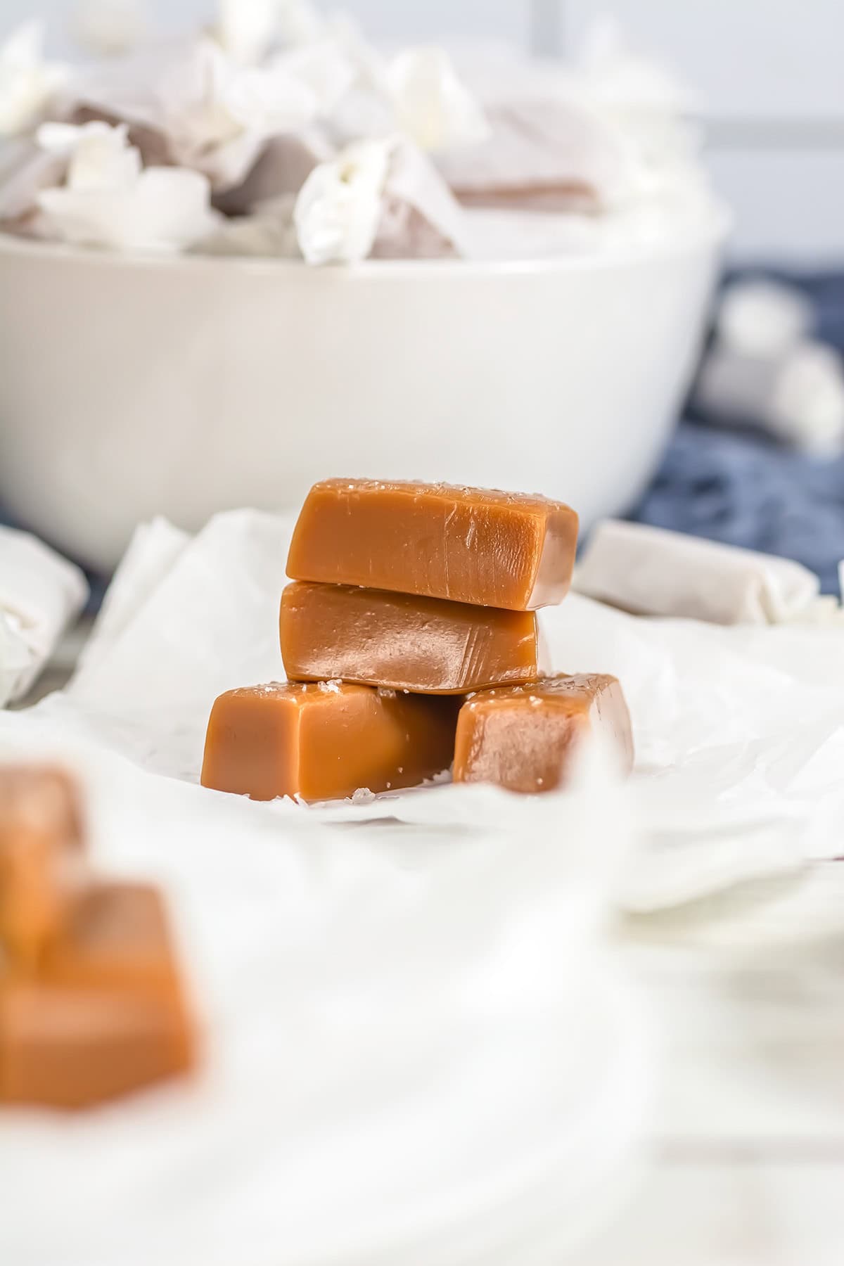 Three stacked salted caramels rest on wax paper, with a bowl of wrapped gourmet sweets in the background.
