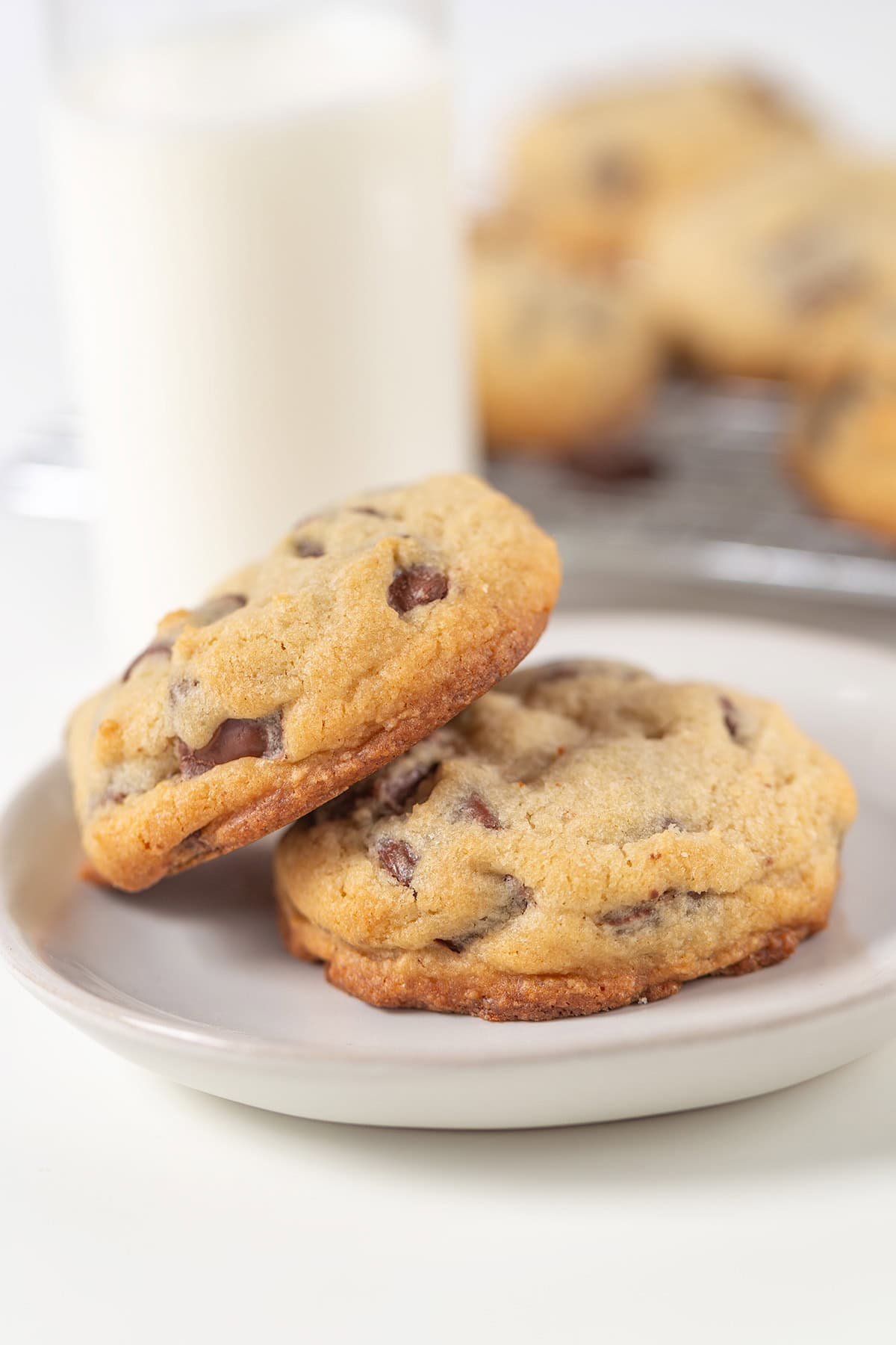 Two Levain Bakery chocolate chip cookies on a gray plate, served with a glass of milk and more cookies in the background.