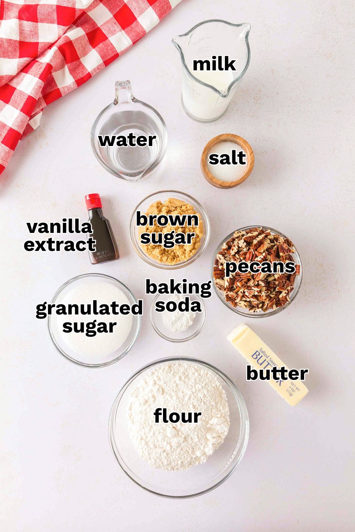 Top-down view of labeled baking ingredients for Pecan Cobbler: milk, water, salt, vanilla extract, brown sugar, pecans, granulated sugar, baking soda, flour, and butter on a white surface.
