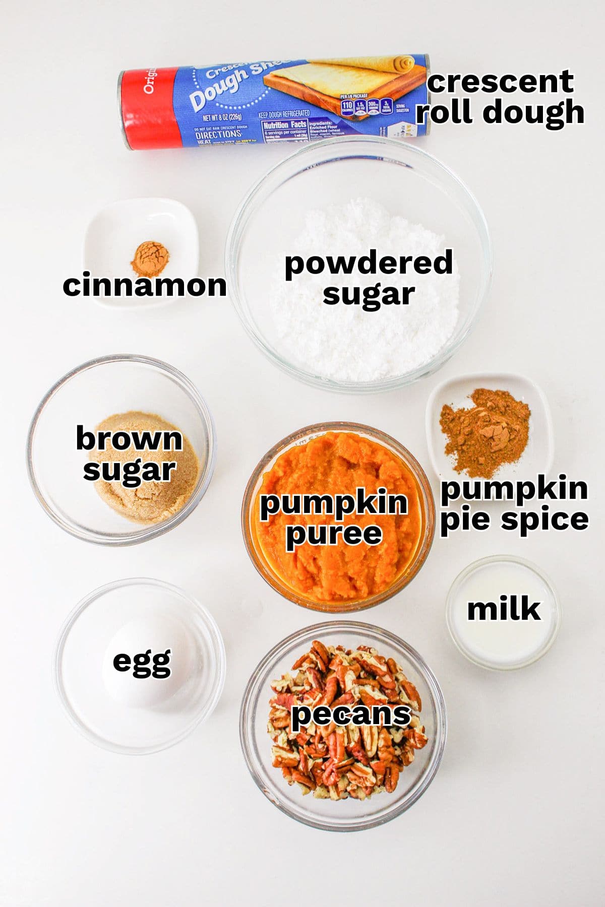 Overhead view of labeled ingredients in bowls for a pumpkin pecan braid: crescent roll dough, powdered sugar, cinnamon, brown sugar, pumpkin puree, pumpkin pie spice, egg, pecans, and milk.