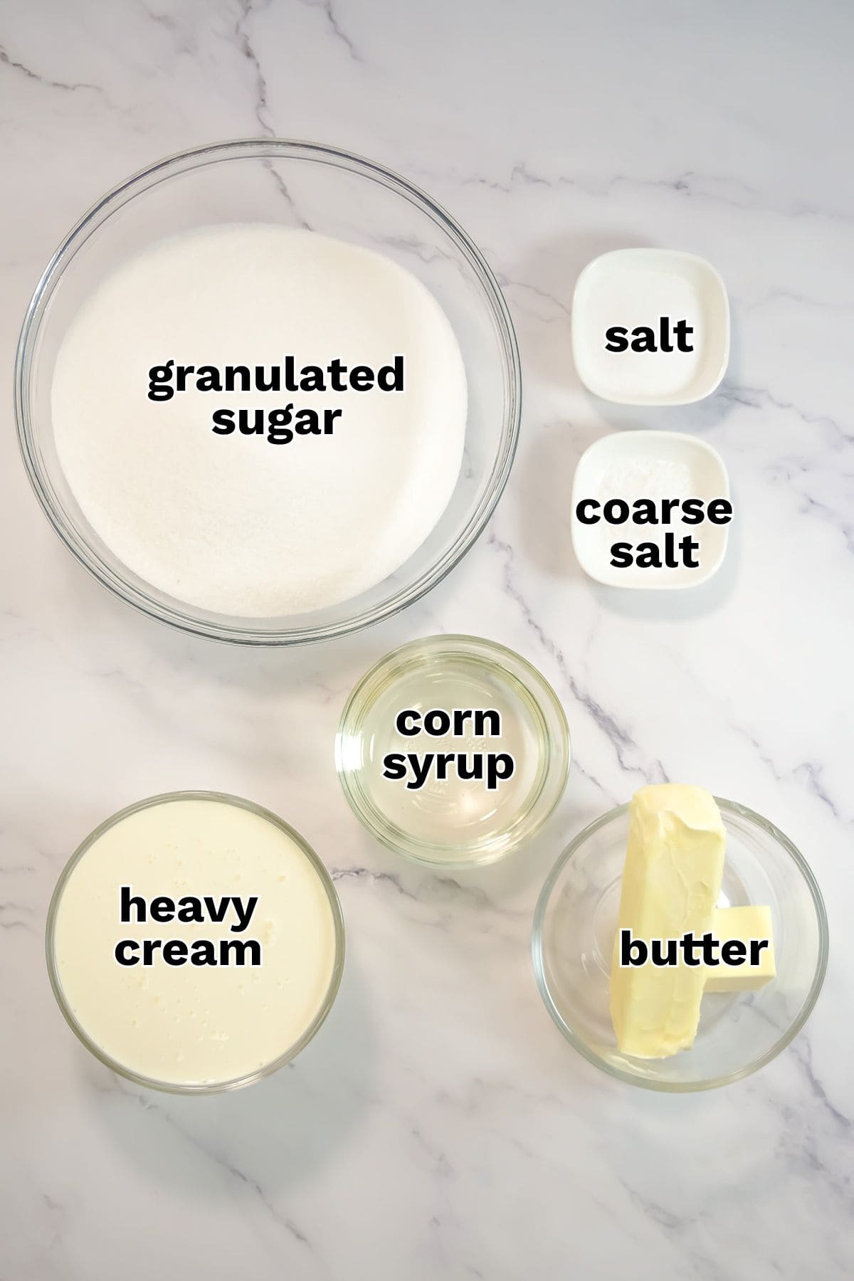 Five labeled bowls on a white surface hold ingredients for gourmet sweets: granulated sugar, heavy cream, corn syrup, coarse salt, regular salt, and a stick of butter—perfect for making rich caramel candy or decadent salted caramels.