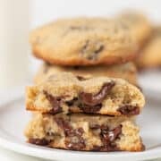 A stack of Levain Bakery Chocolate Chip Cookies, with one broken in half to reveal gooey melted chocolate inside, sits on a white plate.