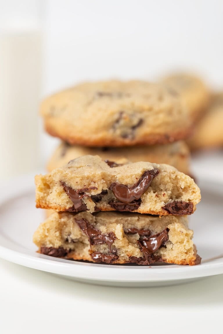 A stack of Levain Bakery Chocolate Chip Cookies, with one broken in half to reveal gooey melted chocolate inside, sits on a white plate.
