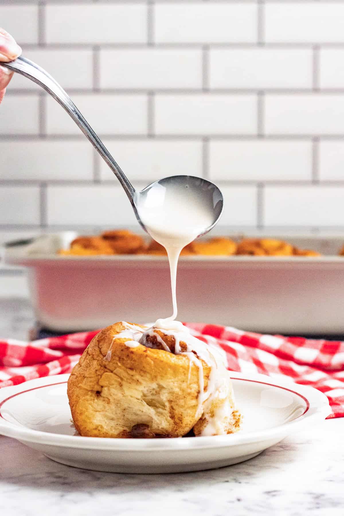 A spoon drizzles white maple icing over a cinnamon roll on a plate, with a pan of cinnamon rolls and a red-and-white towel in the background.