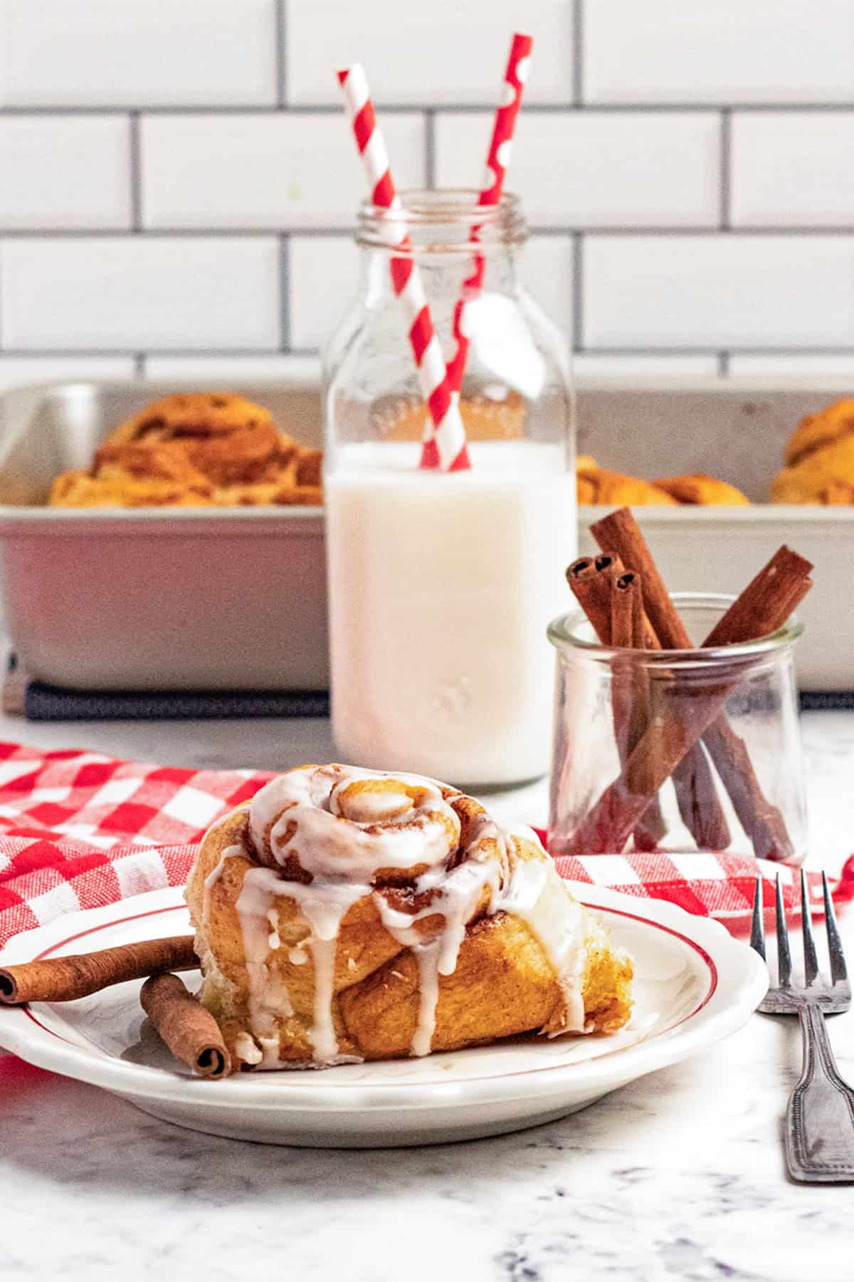 A Maple Cinnamon Roll with icing rests on a plate, garnished with cinnamon sticks, beside a glass bottle of milk with straws and a jar of cinnamon sticks, all set on a table with a red checkered cloth.