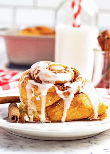 A Maple Cinnamon Roll with icing sits on a white plate beside a cinnamon stick, with a bottle of milk and a red-and-white straw in the background.