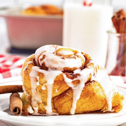 A Maple Cinnamon Roll with icing sits on a white plate beside a cinnamon stick, with a bottle of milk and a red-and-white straw in the background.