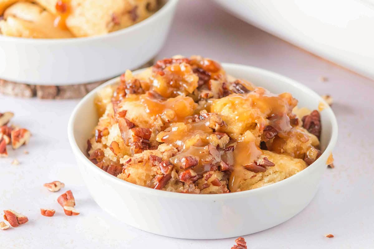 Bowl of sticky bun pieces topped with chopped pecans and caramel sauce, inspired by a classic Southern Recipe, with more dessert pieces and nuts visible in the background.