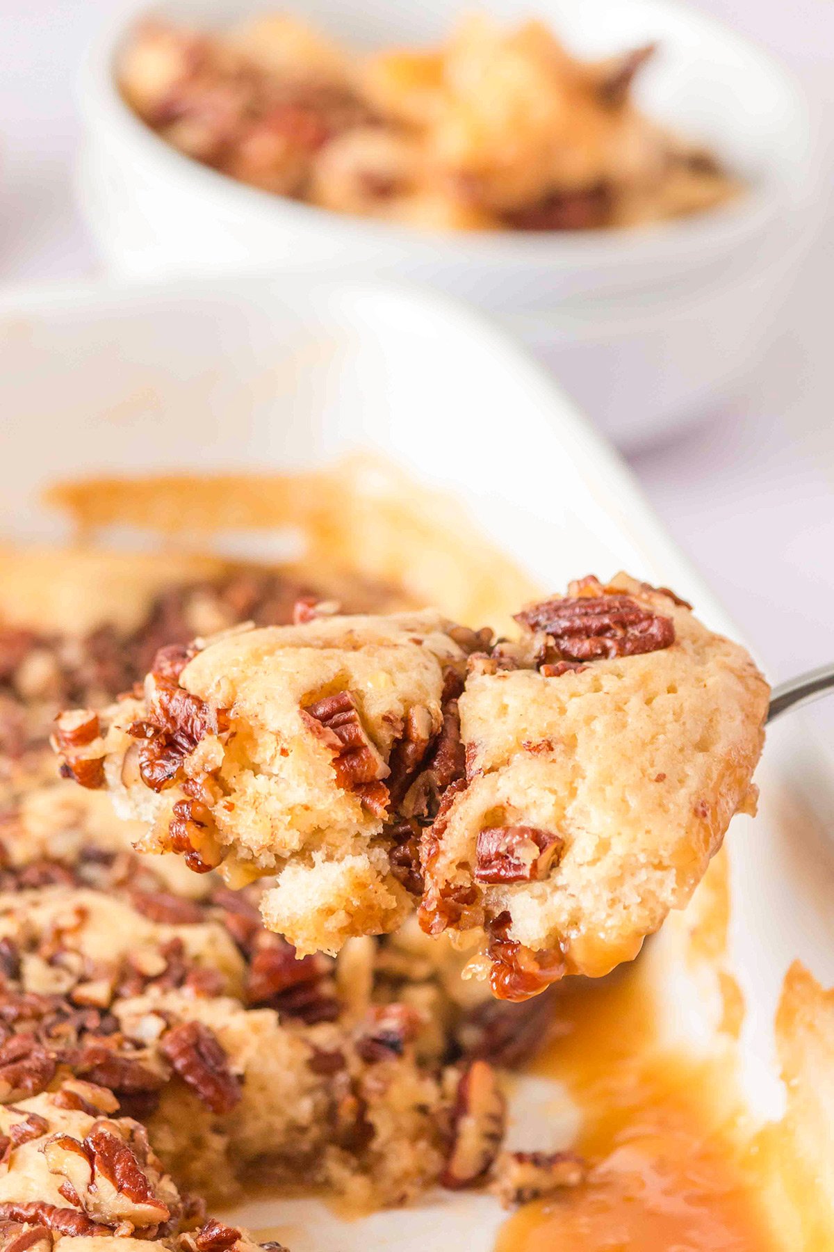 A close-up of a spoon lifting a serving of pecan cobbler from a baking dish, with a bowl of this delicious pecan dessert in the background—perfect for trying out your favorite cobbler recipe.