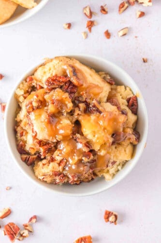 A bowl of Pecan Cobbler topped with chopped pecans and caramel sauce, with scattered pecan pieces on a light-colored surface—a classic Southern Dessert.