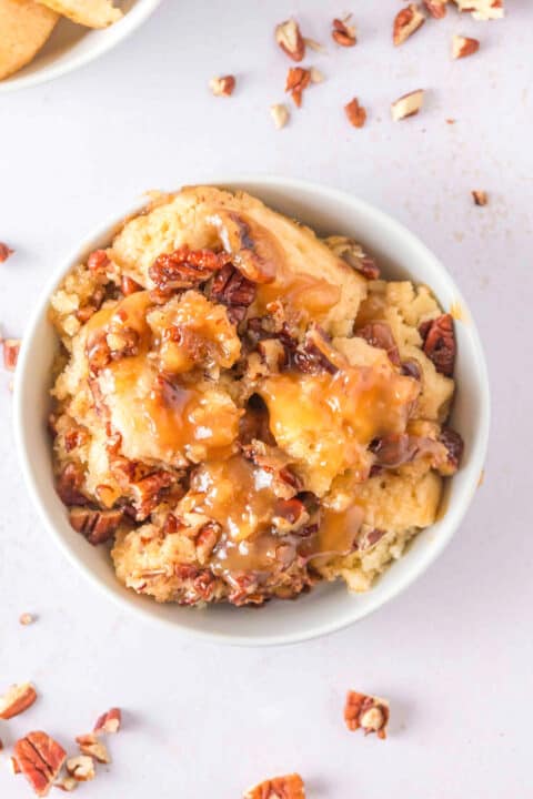 A bowl of Pecan Cobbler topped with chopped pecans and caramel sauce, with scattered pecan pieces on a light-colored surface—a classic Southern Dessert.