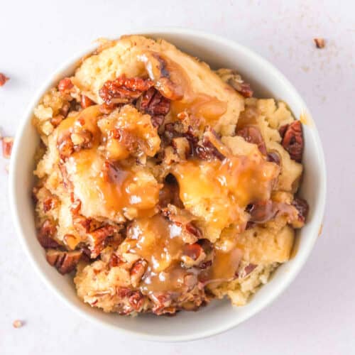 A bowl of Pecan Cobbler topped with chopped pecans and caramel sauce, with scattered pecan pieces on a light-colored surface—a classic Southern Dessert.