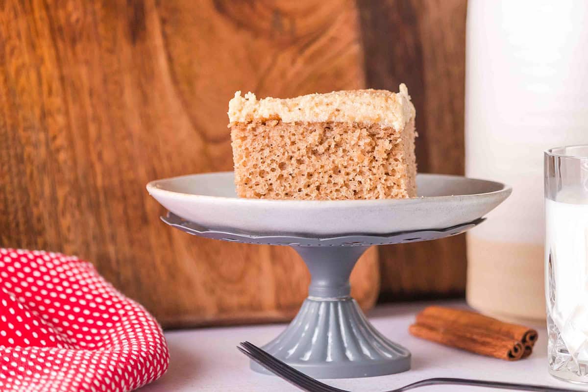 A slice of spice cake with frosting sits on a gray pedestal plate next to a fork, a glass of milk, a cinnamon stick, and a red polka dot cloth—perfect for those seeking an easy spice cake recipe.