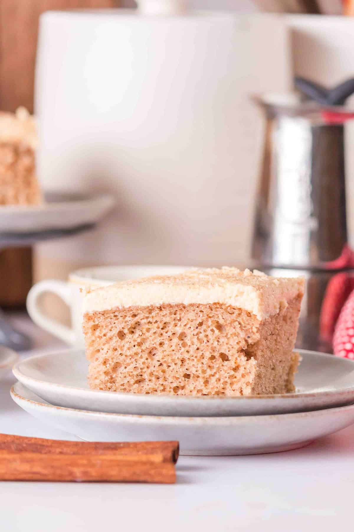 A slice of spice cake sits on a white plate with a cinnamon stick next to it; a cup and metal pitcher are in the background, creating the perfect setting for trying out your favorite spice cake recipe.