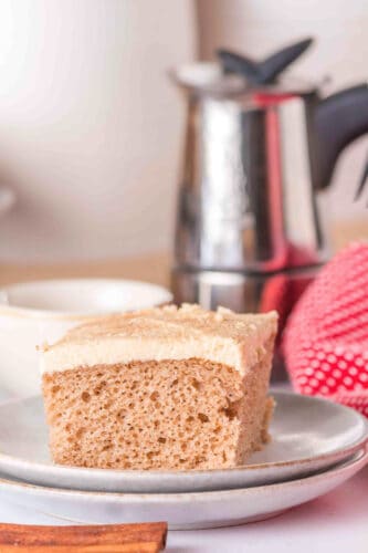 A square piece of frosted spice cake sits on a white plate with a metal coffee pot, a cup, and a red cloth in the background.