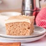 A slice of light brown spice cake sits on a plate, its frosting gleaming, with a blurred background featuring a red towel and kitchen items—a tempting result of a simple spice cake recipe using yellow cake mix.