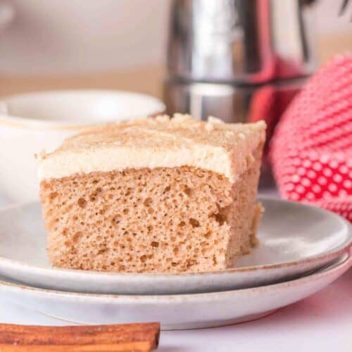 A slice of light brown spice cake sits on a plate, its frosting gleaming, with a blurred background featuring a red towel and kitchen items—a tempting result of a simple spice cake recipe using yellow cake mix.