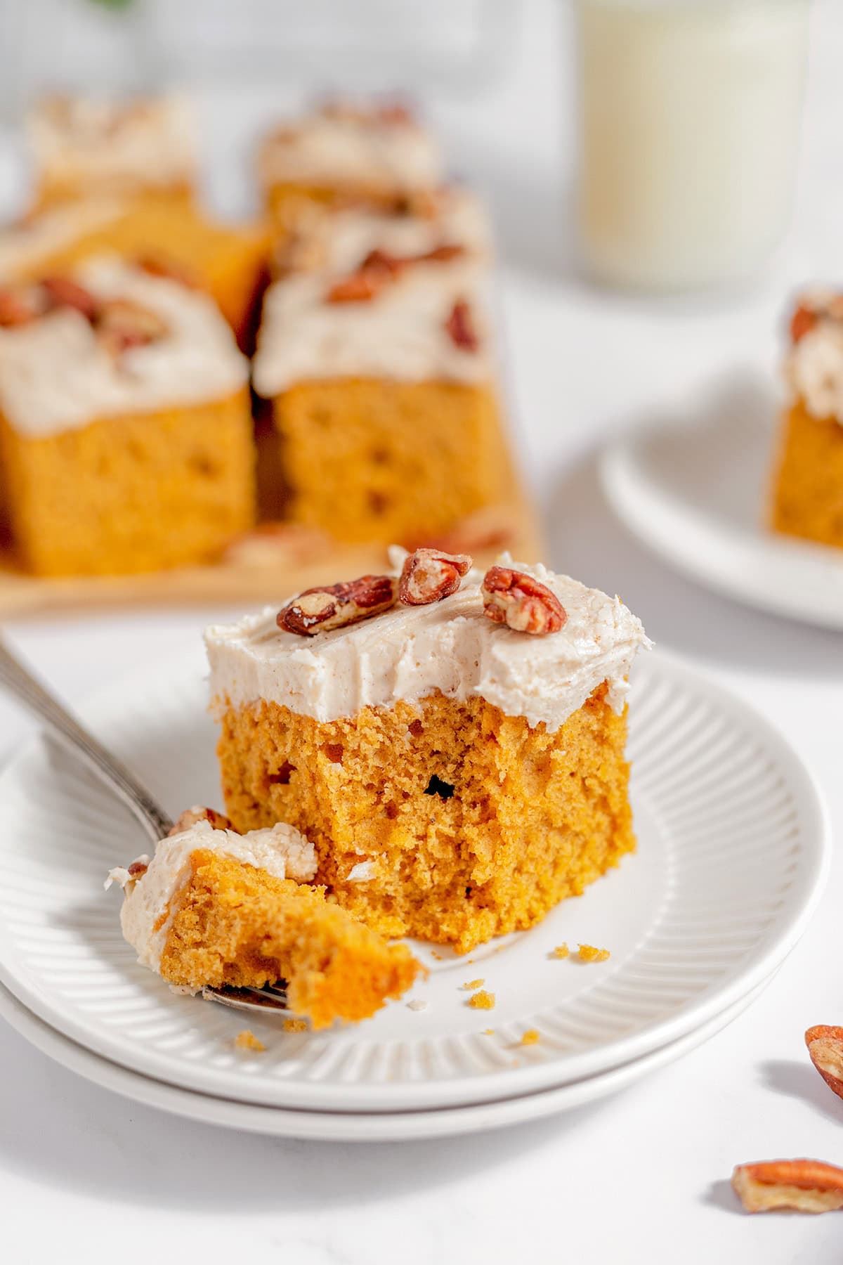 A square slice of sweet potato cake with cream cheese frosting and pecans sits on a white plate, with a fork holding a bite-sized piece. Other cake pieces are in the background.