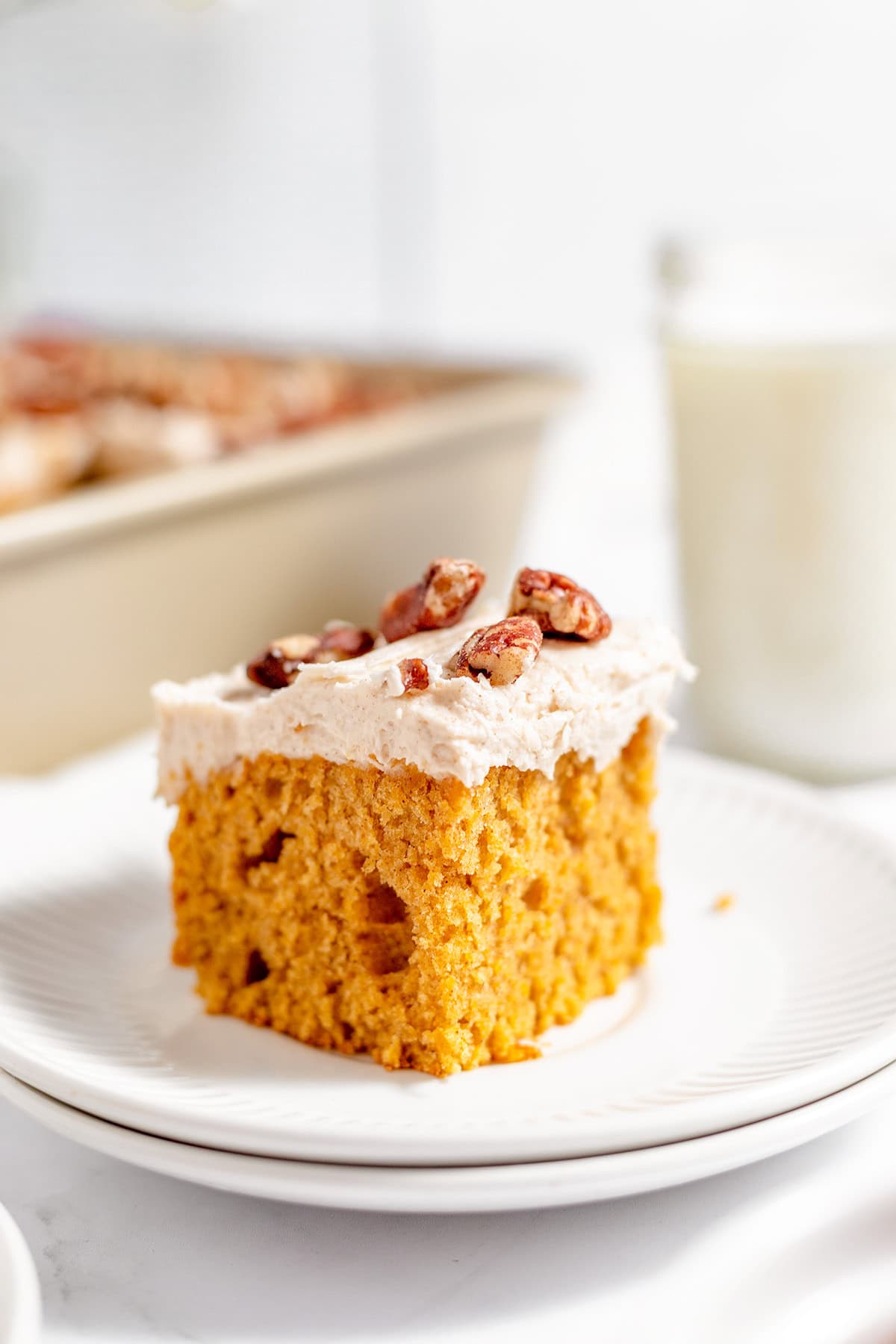 A slice of sweet potato cake with frosting and pecans on top sits on a white plate, with a baking pan and a glass of milk in the background.