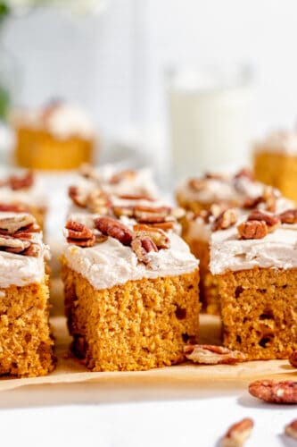 Three rectangular pieces of Sweet Potato Cake topped with frosting and pecan halves are arranged on parchment paper, with more cake slices visible in the background.