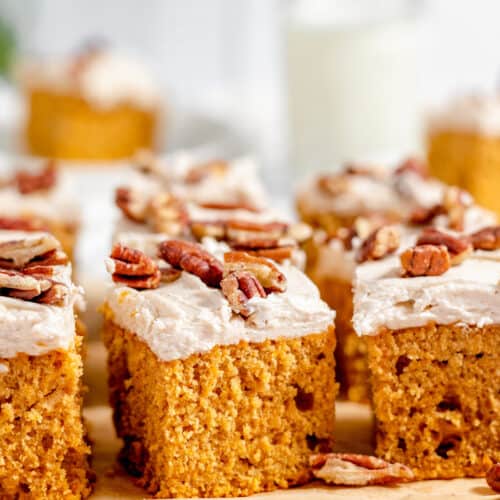 Three rectangular pieces of Sweet Potato Cake topped with frosting and pecan halves are arranged on parchment paper, with more cake slices visible in the background.