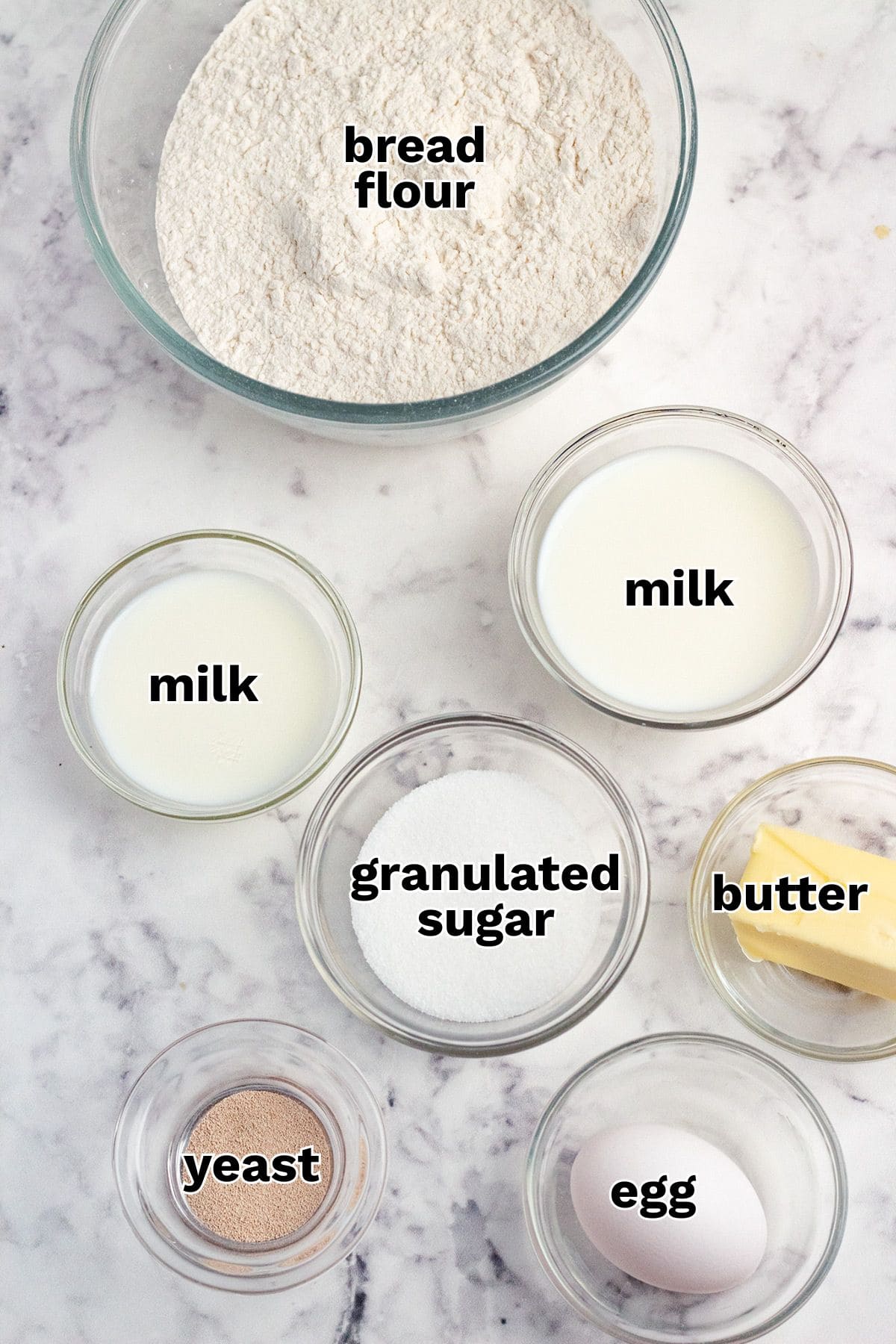 Six labeled bowls on a marble surface contain bread flour, milk, granulated sugar, butter, yeast, and an egg—each ingredient arranged separately for making Maple Cinnamon Rolls.