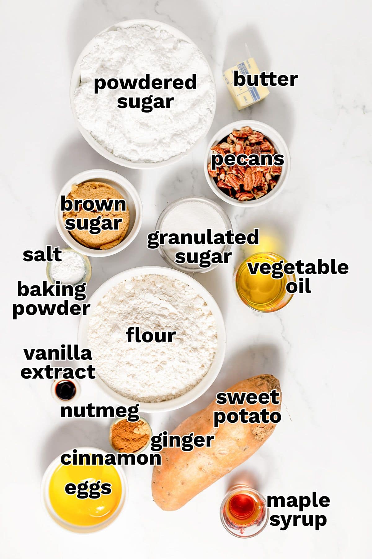 Bowls and containers on a white surface hold ingredients for sweet potato cake, including powdered sugar, brown sugar, granulated sugar, flour, eggs, butter, pecans, vegetable oil, sweet potato, maple syrup, and various spices.