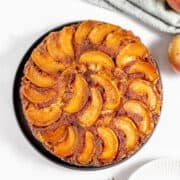 An Apple Upside Down Cake sits elegantly on a black plate, surrounded by fresh apples, a cloth, and two forks, all arranged on a crisp white surface.