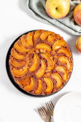 An Apple Upside Down Cake sits elegantly on a black plate, surrounded by fresh apples, a cloth, and two forks, all arranged on a crisp white surface.
