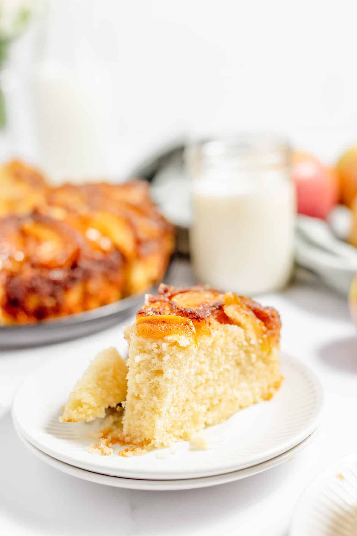 A slice of pineapple upside-down cake on a white plate sits beside a glass of milk, with the rest of the cake in the background—perfect for anyone who also enjoys Apple Upside Down Cake.