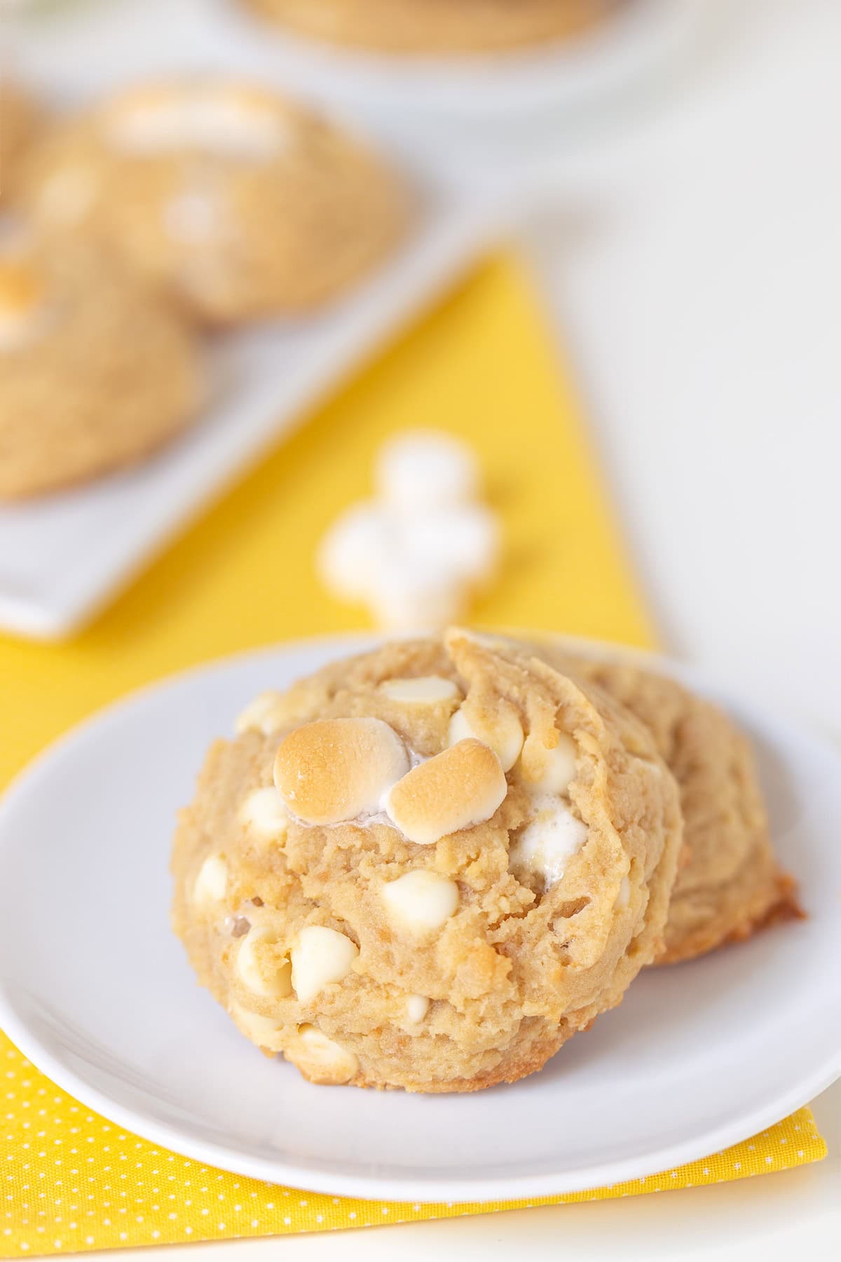 Two banana pudding cookies with white chocolate chips and mini marshmallows are placed on a white plate, set on a yellow napkin.