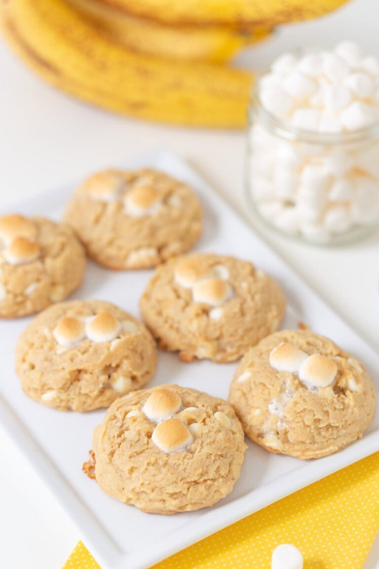 Six banana pudding cookies topped with mini marshmallows are arranged on a white rectangular plate, with a jar of marshmallows and bananas in the background.