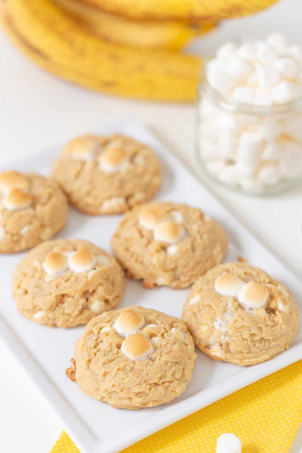 Six banana pudding cookies topped with mini marshmallows are arranged on a white rectangular plate, with a jar of marshmallows and bananas in the background.
